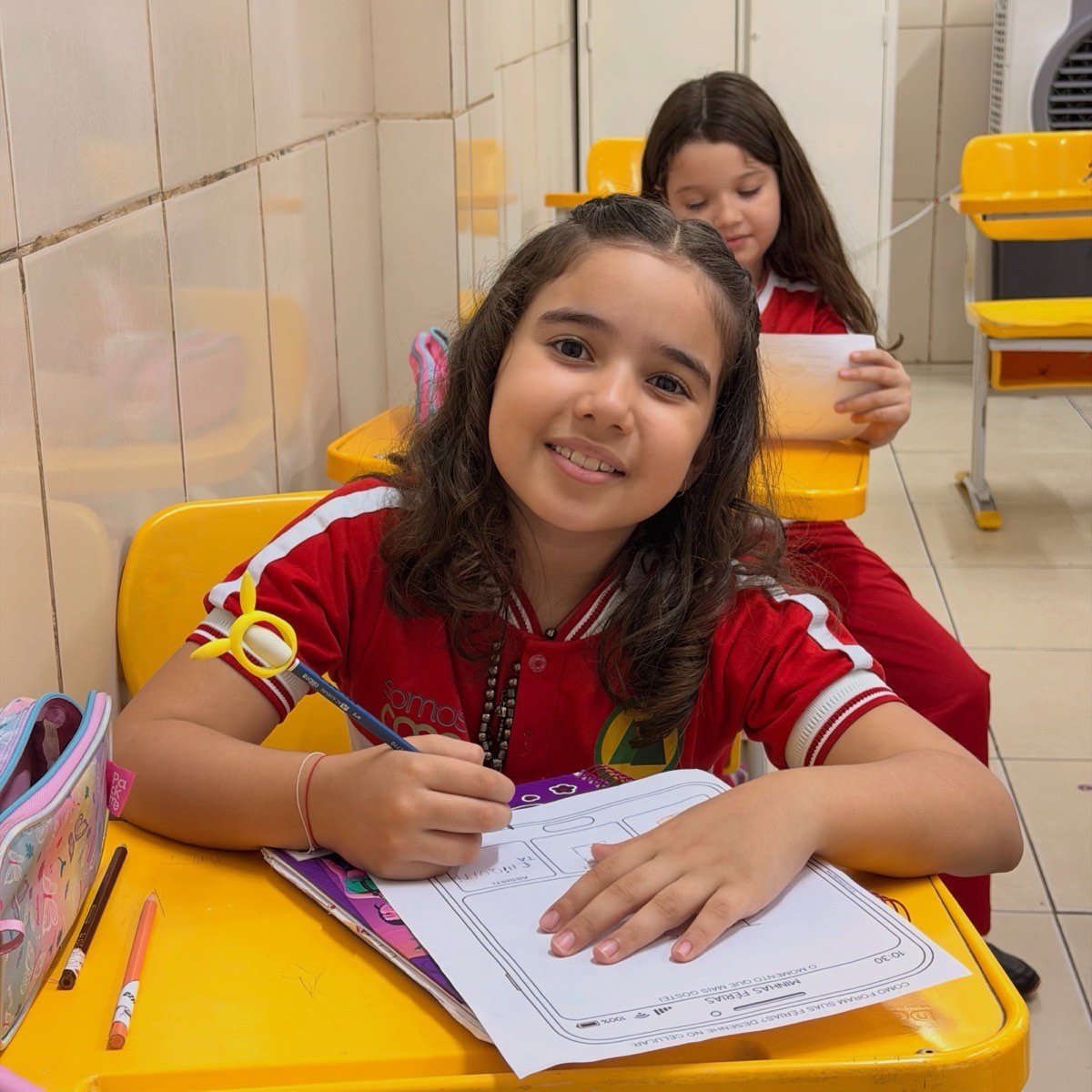 Aluna da CEDEF sorrindo enquanto faz uma atividade na carteira, de uniforme vermelho da escola